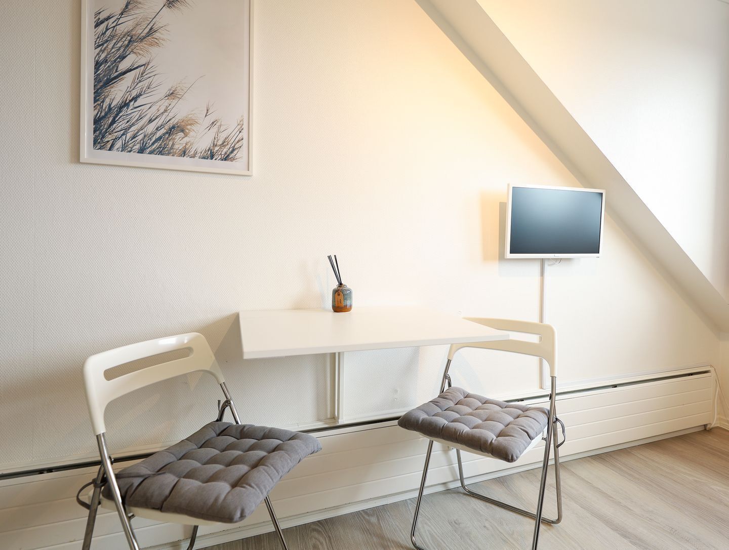 A modern room with a white desk, two cushioned chairs, a small vase, and a mounted TV.