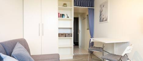 A compact living space with a grey sofa, a white desk, and a shelving unit with books and baskets.