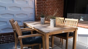 Une table en bois placée à l’extérieur, avec deux chaises, une plante en pot et un mur de briques en arrière-plan.
