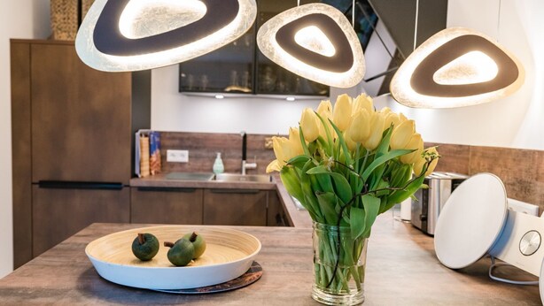 Une cuisine moderne avec des luminaires suspendus, un panier de fruits et un vase de fleurs posé sur le plan de travail.