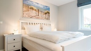 A bedroom with a bed, a nightstand, a drawer, a window with a view of the beach, and a painting of a beach scene. - Strandhütte - House Strandhütte (Sylt)