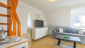 A modern living room with a wooden staircase, a white sofa, a flat-screen TV, and a dining area with a table and chairs.