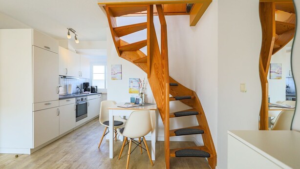 A modern kitchen with white cabinets, a dining area with a table and chairs, and a wooden staircase.