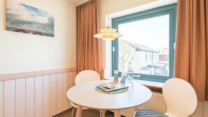 Et værelse med et rundt bord, to stole, et vindue med gardiner, en hængende lampe og et billede på væggen.