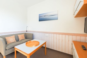 A small, cozy room with a grey sofa, a wooden table with a tray, a painting of a calm body of water on the wall, and a TV mounted on the wall.