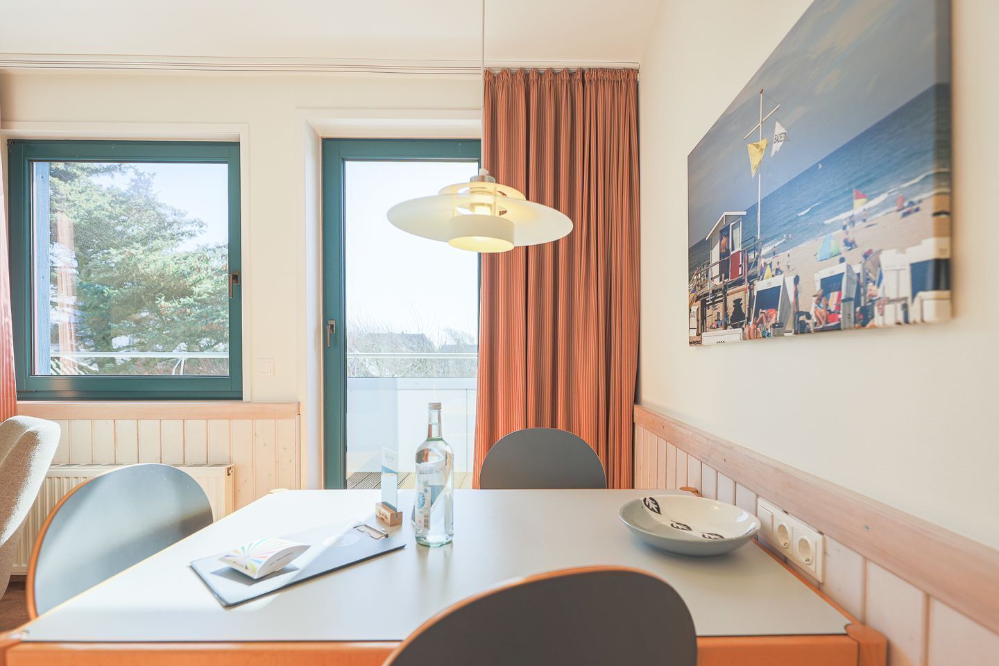Una habitación de hotel con una mesa de comedor, sillas, una lámpara colgante, una ventana con cortinas y un cuadro colgado en la pared.