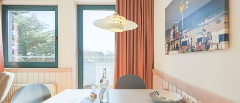 Una habitación de hotel con una mesa de comedor, sillas, una lámpara colgante, una ventana con cortinas y un cuadro colgado en la pared.