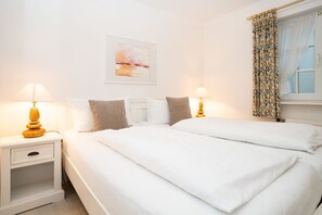Un lit double avec du linge de lit blanc, deux tables de chevet avec des lampes, une œuvre d’art encadrée au-dessus du lit et une fenêtre avec des rideaux à motifs.