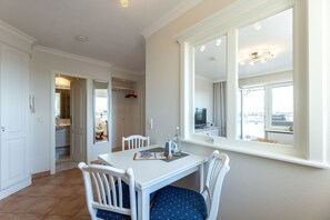 Un coin repas avec une table et des chaises blanches, un grand miroir qui reflète la pièce et une fenêtre donnant sur l’extérieur.