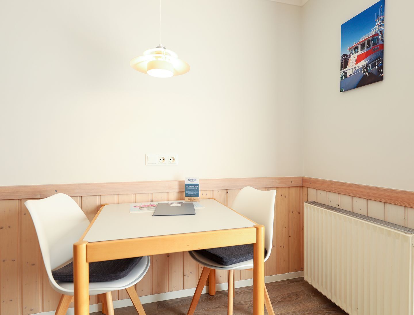 A small dining area with a wooden table and two white chairs, a pendant light, and a framed picture on the wall.