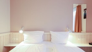 Ein Doppelbett mit weißen Bettlaken, zwei Nachttischlampen, ein Spiegel und ein brauner Vorhang sowie ein hölzernes Kopfteil.