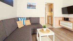 Ein modernes Wohnzimmer mit einer grauen Eckcouch, einem kleinen weißen Beistelltisch mit einer Pflanze und einem an der Wand montierten Flachbildfernseher.