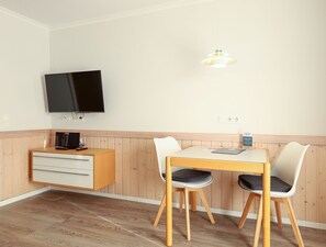 Una habitación pequeña y ordenada con un armario de madera, un televisor montado en la pared, una mesa con dos sillas y una lámpara colgante.