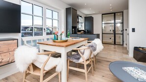 A modern kitchen with a dining area, wooden chairs with fluffy cushions, a wooden table, and a view of parked cars outside. - Hüs gur seen, Germany (Sylt)