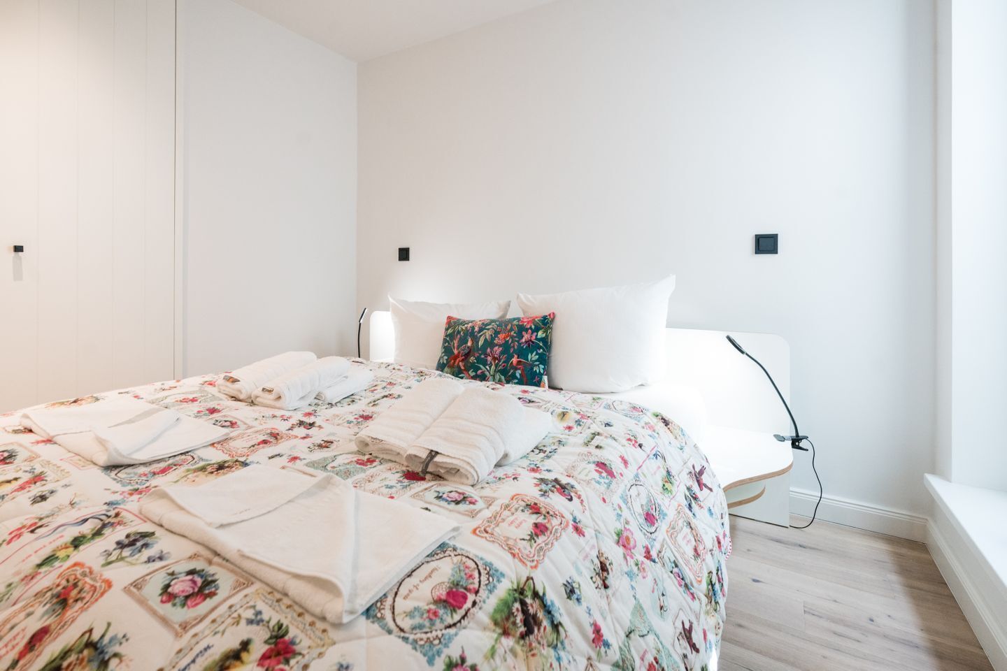 Una camera da letto moderna con un letto grande, biancheria da letto floreale, pareti bianche e un comodino con una lampada.