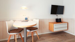 A small, modern room with a round dining table, two chairs, a television mounted on the wall, and a wooden cabinet with drawers.