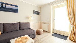 A modern living room with a grey sofa, a small white table with a woven placemat, a floor lamp, and a large window with curtains.