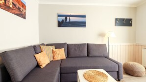 A modern living room with a grey sectional sofa, a small white side table, a woven placemat, a lamp, and a wall-mounted coat rack.
