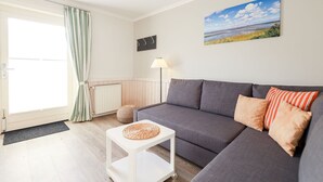 Un salon avec un canapé gris, une petite table d’appoint blanche, une lampe et un tableau représentant un paysage accroché au mur.