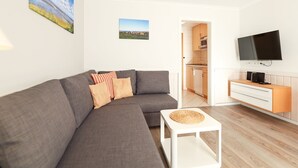 Un salon moderne avec un canapé d’angle gris, une petite table d’appoint blanche, un téléviseur à écran plat fixé au mur et un coin cuisine visible à travers une porte ouverte.