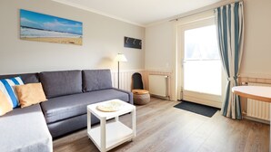 A modern living room with a grey sofa, a small white side table, a lamp, and a view of the outdoors through a large window.