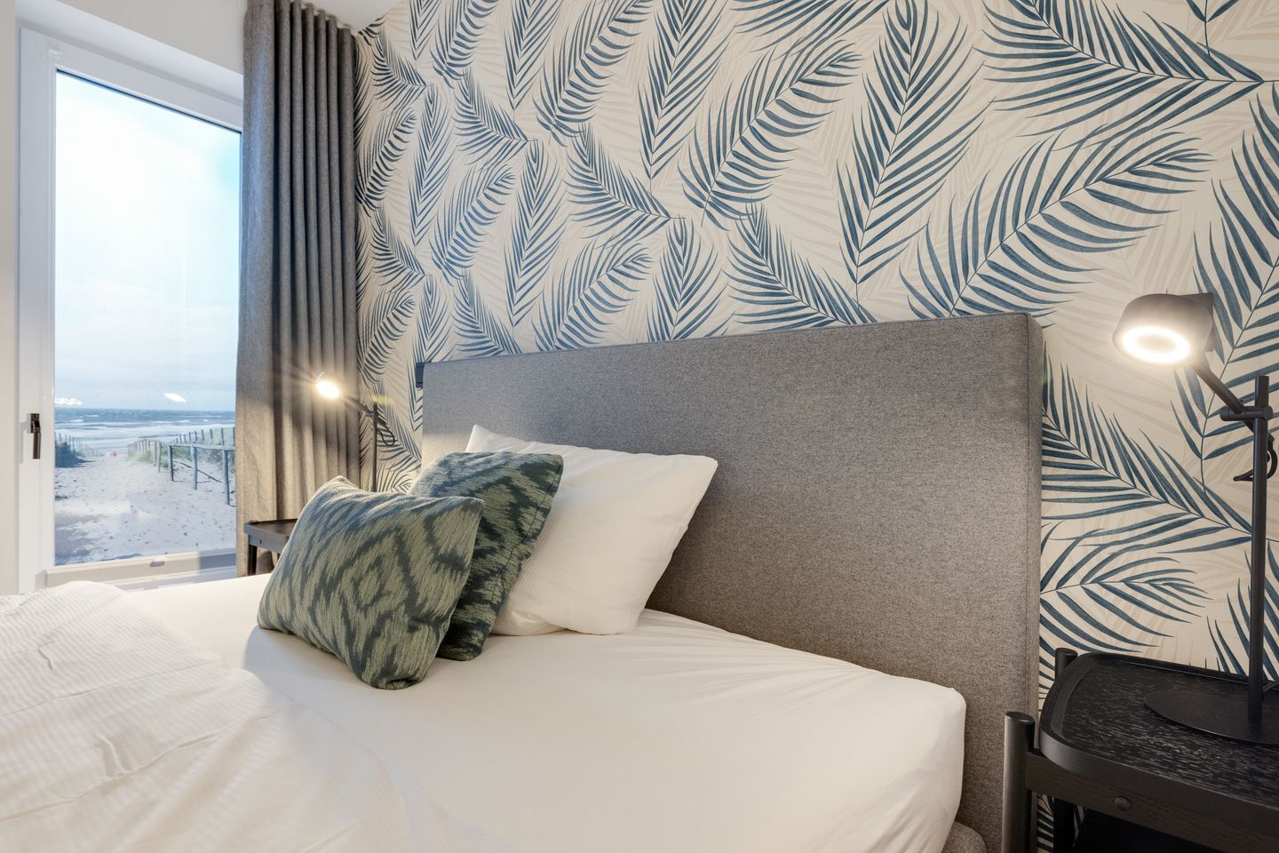 A hotel room with a bed, a headboard, a nightstand, a lamp, and a view of the beach.