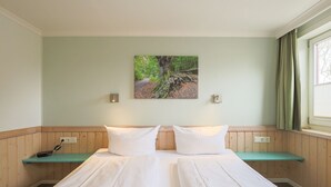 A double bed with two pillows, a painting of a forest on the wall, and a window with curtains. - Imke - House Rolf - Imke (Sylt)