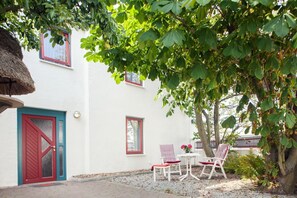 A white building with a red door and windows, a thatched roof, and outdoor seating with red and white striped cushions. - Imke - House Rolf - Imke (Sylt)
