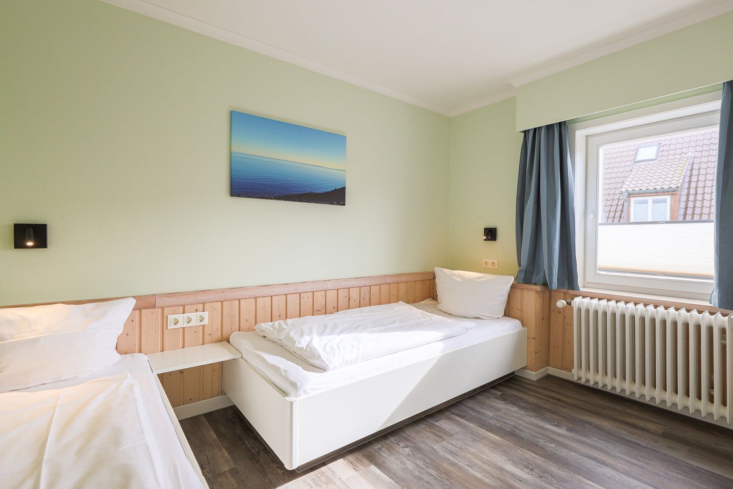 Une chambre avec un lit simple, une table de chevet, une lampe fixée au mur, une fenêtre avec des rideaux et un radiateur.