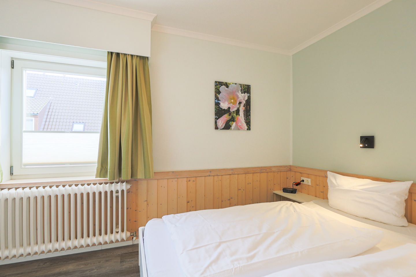 Ett hotellrum med en säng, ett fönster med gardiner, ett element, en vägglampa och en tavla.