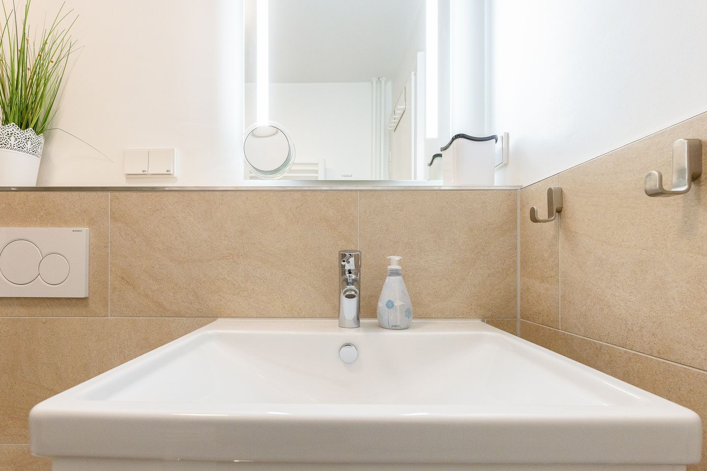A bathroom with a white sink, a mirror, a plant, and a soap dispenser.