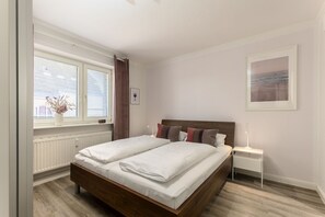Un dormitorio con cama de madera, ropa de cama blanca, una mesita de noche, una lámpara, una ventana con persianas, un radiador y un cuadro en la pared.