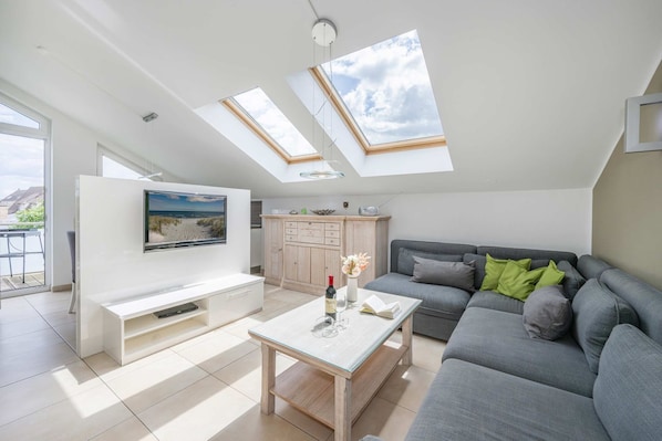 A modern living room with a flat-screen TV, a grey sofa, a coffee table, and a dining area with a white cabinet.