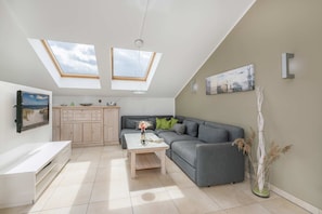 A modern living room with a grey sofa, a coffee table, a TV, and a kitchen area with wooden cabinets. There are skylights above the sofa and a painting on the wall.