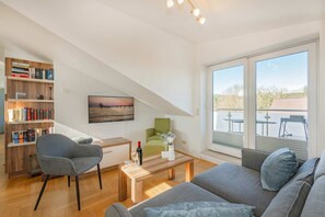 Una sala de estar moderna con un sofá, una butaca, una mesa de centro, una estantería, un televisor y un balcón con mobiliario exterior.