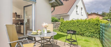 A patio with a table and chairs, a grill, and a view of a house with a white exterior and a red roof.