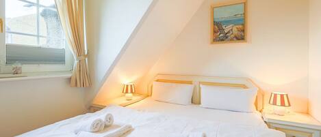Una cama bien hecha con sábanas y toallas blancas, dos lámparas de noche, un cuadro de una playa y una ventana con cortinas.
