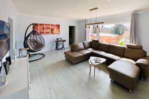 Un salon moderne avec un canapé d’angle, un fauteuil suspendu, une machine à coudre et une grande fenêtre donnant sur un beau paysage.