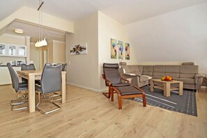 Un salon moderne avec un canapé, des fauteuils, une table à manger avec des chaises, et un coin cuisine à l’arrière-plan.