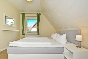 Ein Schlafzimmer mit Bett, Nachttisch, Lampe, Fenster mit Vorhängen und Blick ins Freie.