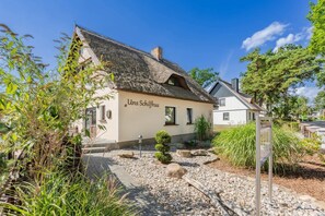 Un bâtiment traditionnel avec un toit de chaume, une enseigne indiquant « Unser Schloss », et un jardin bien entretenu avec des allées de gravier et de la verdure.
