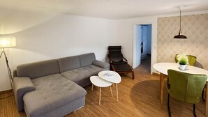 Ein modernes Wohnzimmer mit einem grauen Ecksofa, einem kleinen weißen Tisch, einem schwarzen Ledersessel und einem Essbereich mit einem grünen Stuhl.