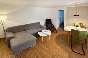 Un salon moderne avec un canapé d’angle gris, une petite table blanche, un fauteuil en cuir noir et un coin repas avec une chaise verte.