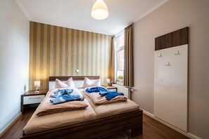 Uma cama de casal com cabeceira de madeira, duas mesas de cabeceira com abajures, uma parede listrada, uma janela com cortinas e um cabideiro fixado na parede.