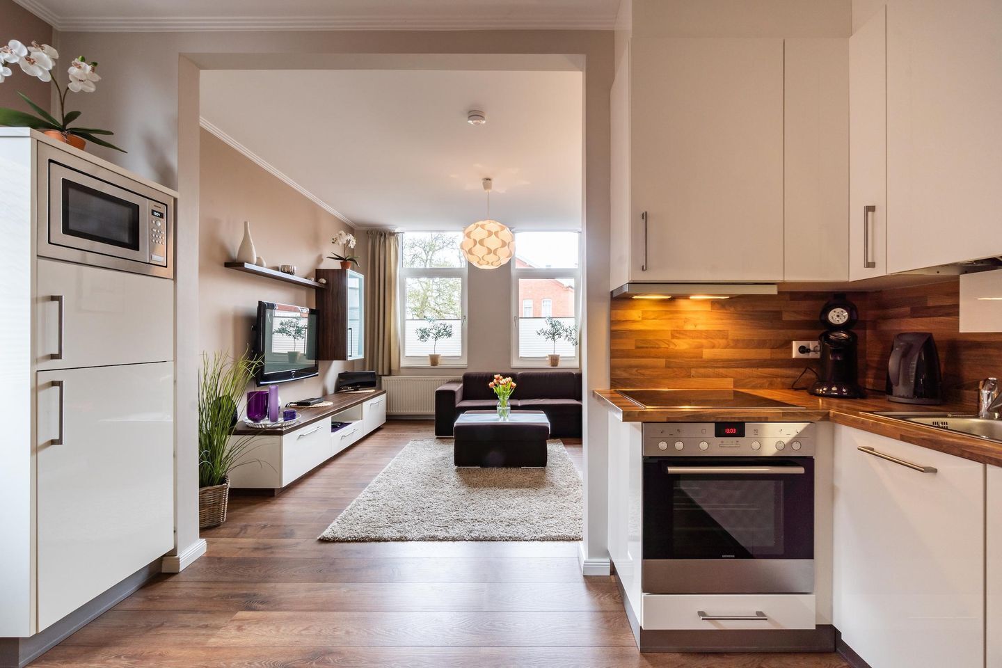 Una cocina moderna con horno integrado, microondas y cafetera. Hay una zona de comedor con sofá y televisor.