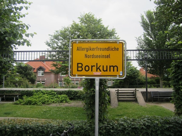 Un panneau jaune avec une inscription noire indiquant « Île de la mer du Nord adaptée aux personnes allergiques : Borkum » est placé devant un bâtiment avec un toit rouge et une clôture.