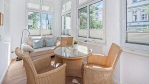 A bright room with a round glass table surrounded by wicker chairs, a sofa with cushions, and large windows with blinds.