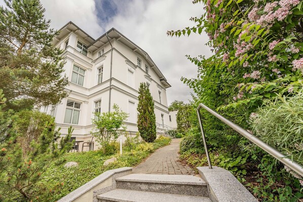 A building with a staircase leading to a door, surrounded by greenery and a metal railing.