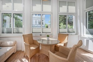 Ein sonnendurchfluteter Raum mit einem runden Glastisch, umgeben von Korbstühlen, großen Fenstern mit Jalousien und Blick auf Gebäude im Freien.