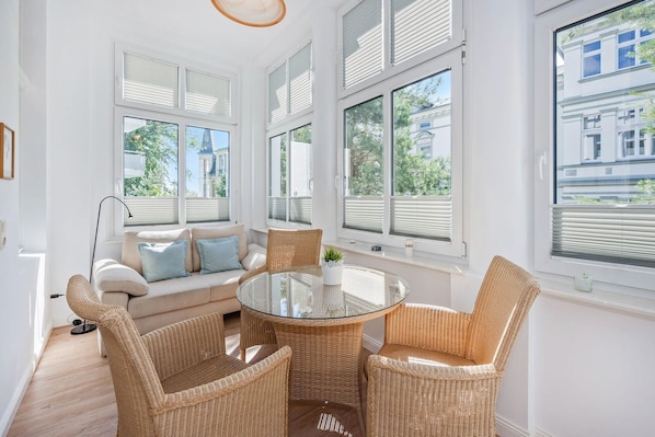 A bright room with a round glass table surrounded by wicker chairs, a sofa with cushions, and large windows with blinds.